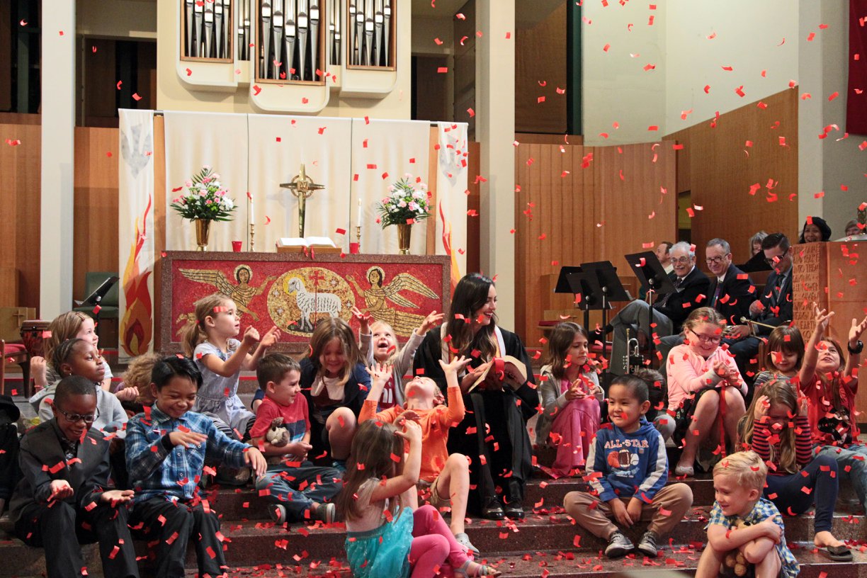 Pastor Jen sitting on the altar steps with a large group of children, laughing and looking up as red confetti floats down on top of them.