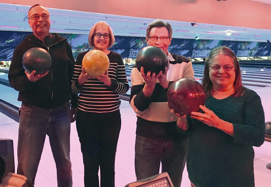Four adults at the bowling alley, holding bowling balls and smiling at the camera