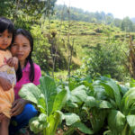 A woman and child kneel and look at the camera amidst a growing garden