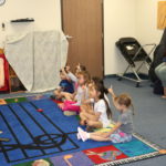 A group of young children sit on the floor and watch their music instructor