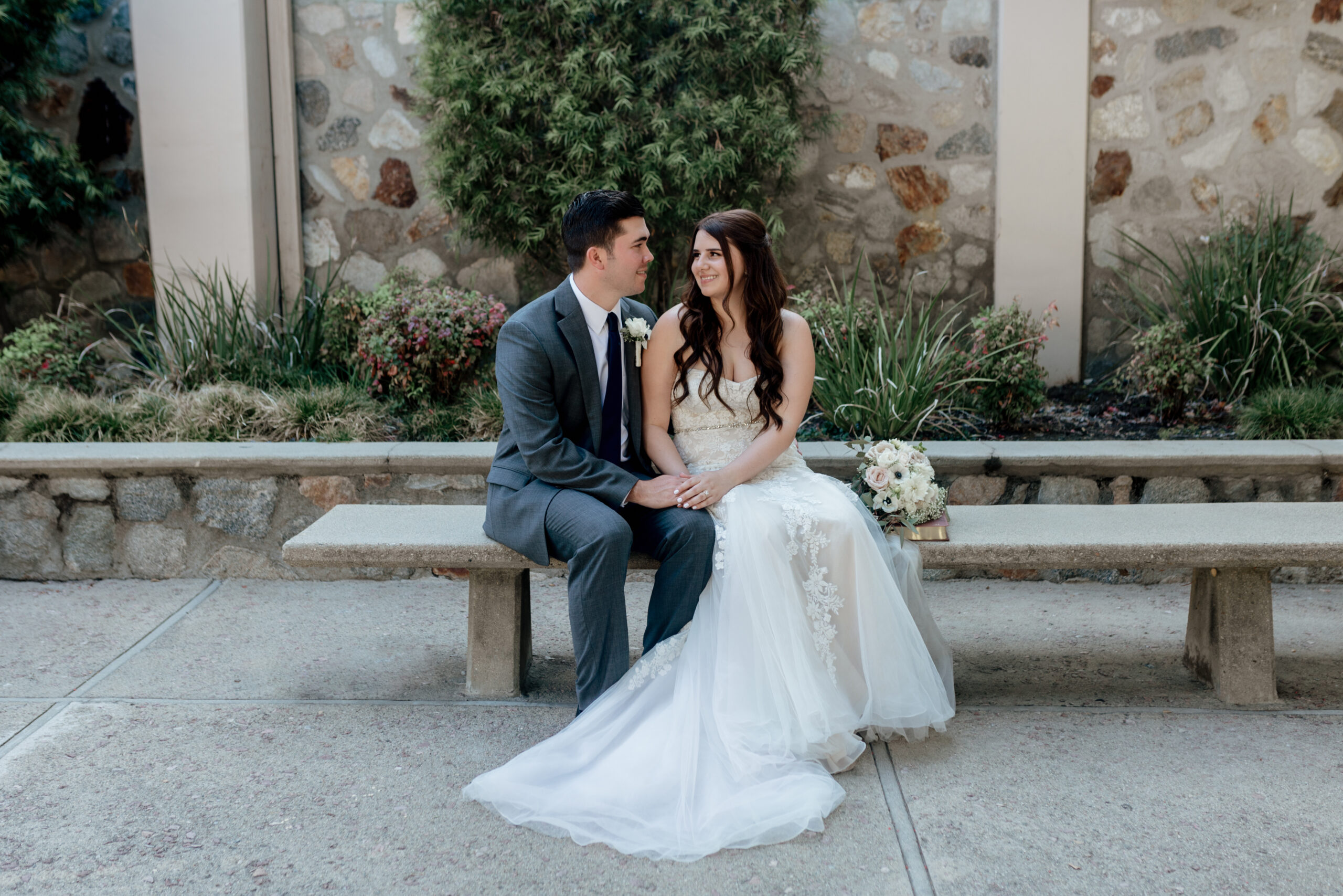 Two newlyweds, looking at each other and sitting on a bench in front of green plants