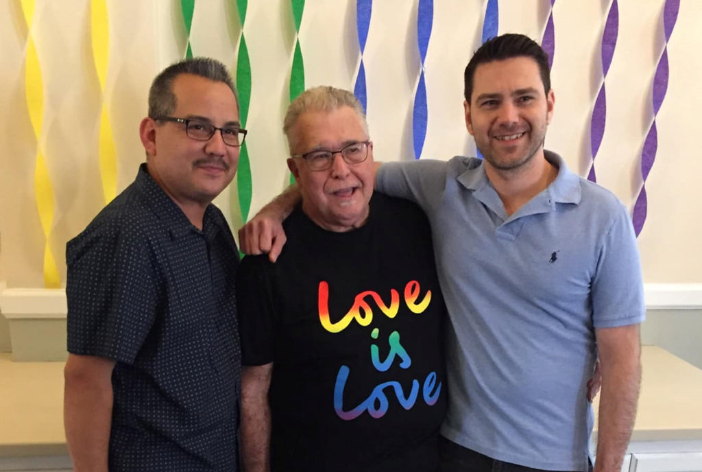 Three men smiling with their arms around each other, with rainbow decorations behind them.